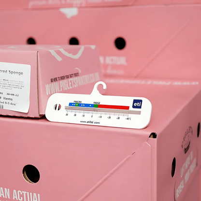 Thermomètre numérique accroché à une boîte en carton rose, affichant une lecture de température, placé sur d'autres boîtes roses en arrière-plan. Ce Thermomètre horizontal pour réfrigérateur et congélateur de Thermomètre.fr est conçu.